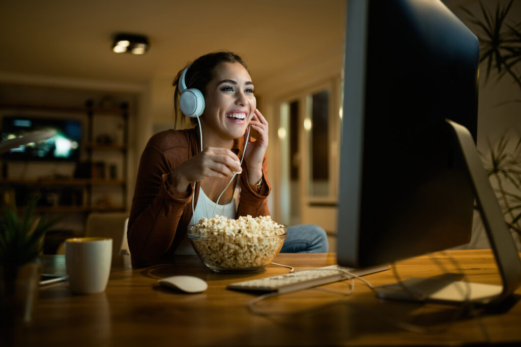 Happy woman eating popcorn and watching movie on desktop PC while enjoying at night at her apartment.